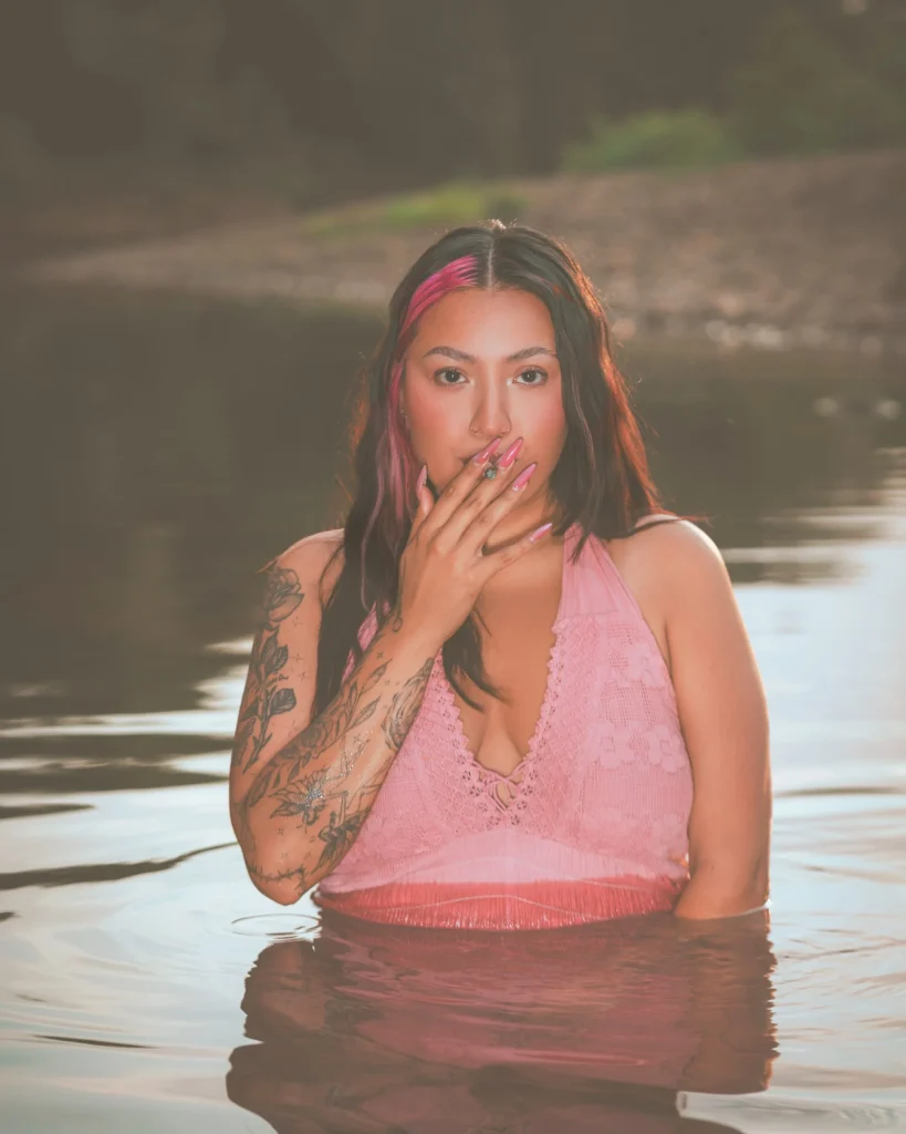 Model posing in water with tattoos and pink hair accents.