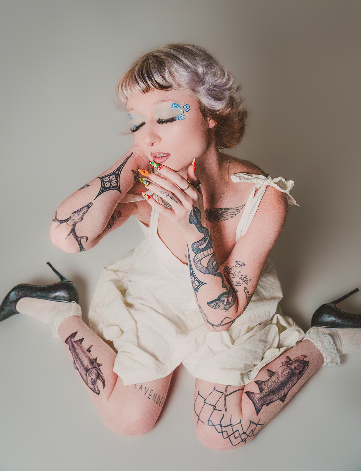 Tattooed model poses in stylish outfit with striking makeup.